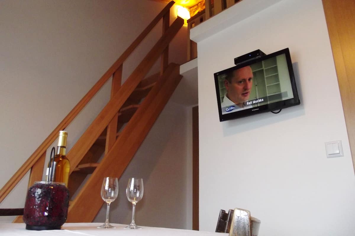 A room with a wooden staircase, a television mounted on the wall, and a table with a bottle and two glasses.