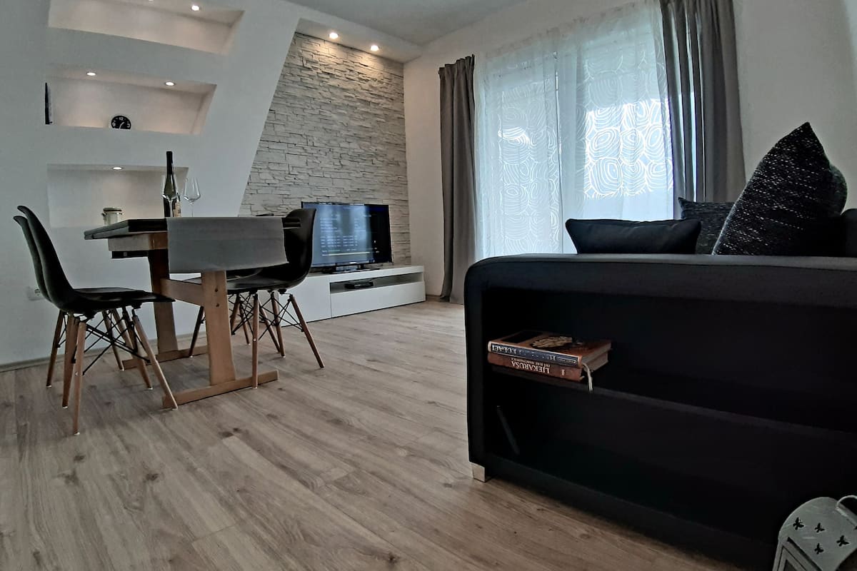 A modern living room with a black sofa, a dining table with chairs, a TV, and a bookshelf.