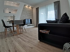 Una sala de estar moderna con un sofá negro, una mesa de comedor con sillas, un televisor y una estantería.