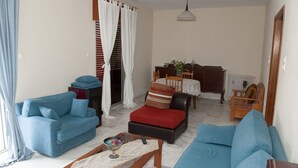 Una sala de estar con un sofá azul, un reposapiés rojo y una mesa de centro de madera. En el fondo, se ve una mesa de comedor con sillas.