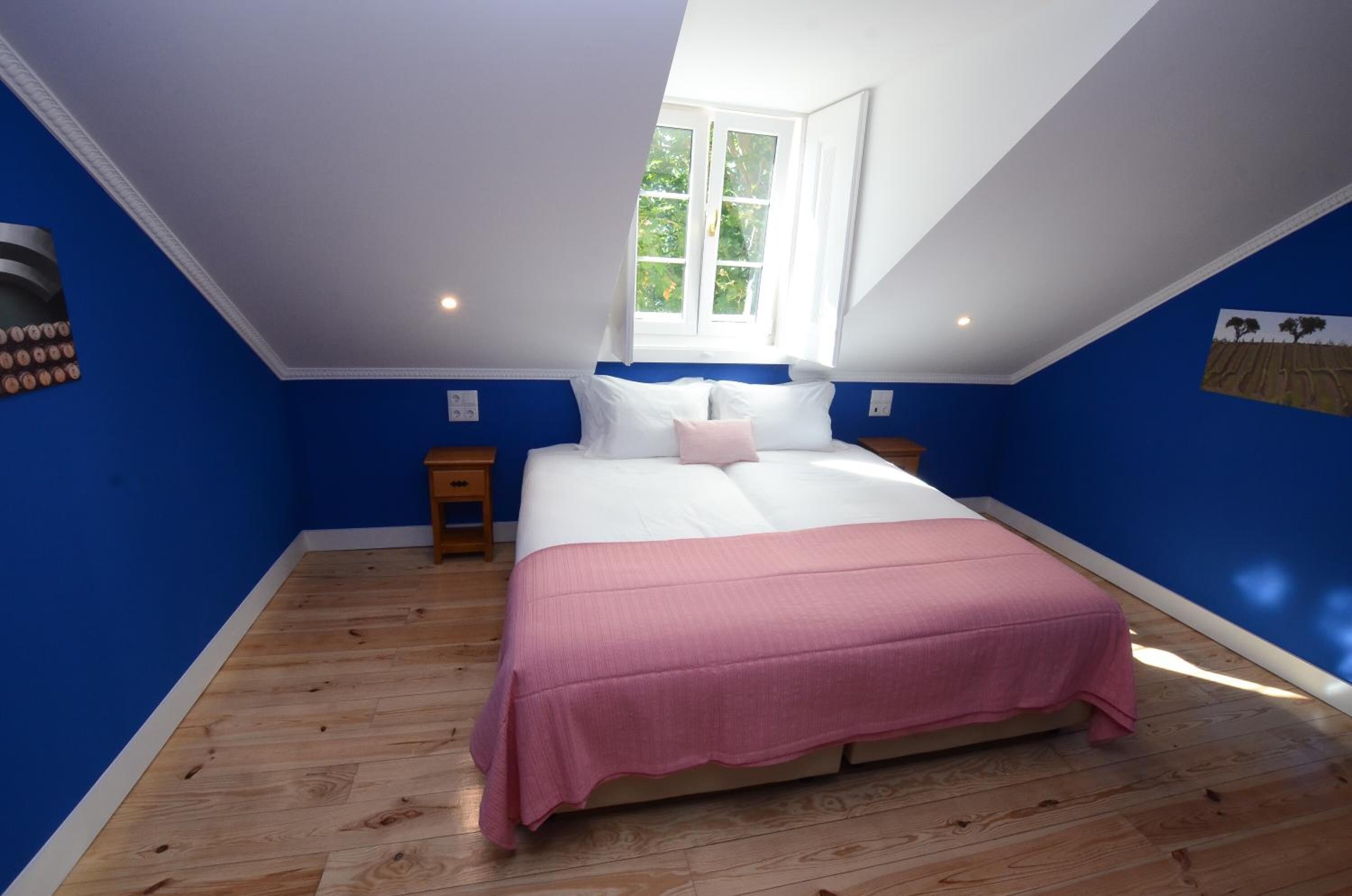 A bedroom with a sloped ceiling, a large bed with a pink bedspread, a window with white curtains, and a wall-mounted clock.