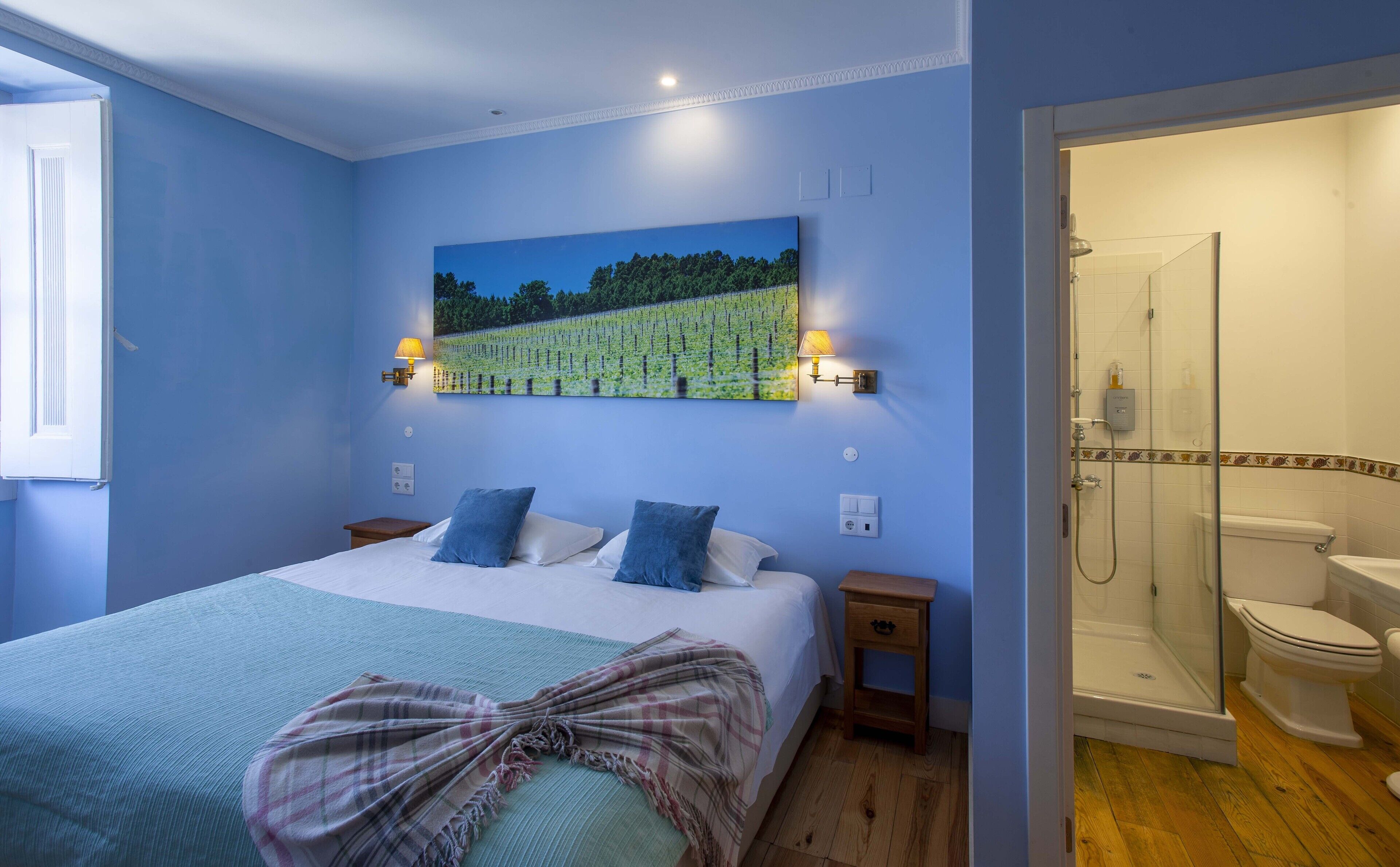 A bedroom with a large bed, a painting of a vineyard, bedside lamps, and a glass-enclosed shower.