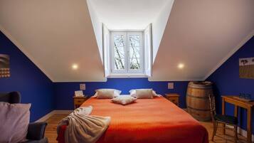 Une chambre avec un plafond en pente, un grand lit recouvert d’une couverture orange, un tonneau en bois et une petite table avec des verres.
