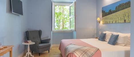 Un dormitorio con cama, una silla, una mesita, una ventana y un cuadro en la pared.