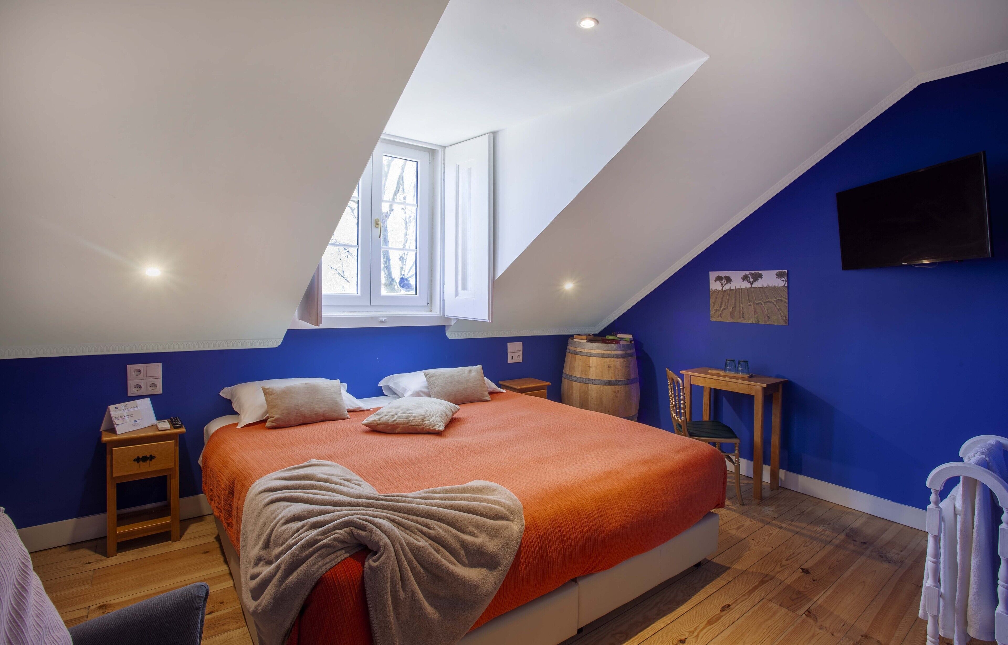 Ein Schlafzimmer mit schrägem Dach, einem großen Bett mit orangefarbener Bettdecke, einem Holz-Nachttisch, einem kleinen Tisch mit Fassform und einem Wand-Fernseher.