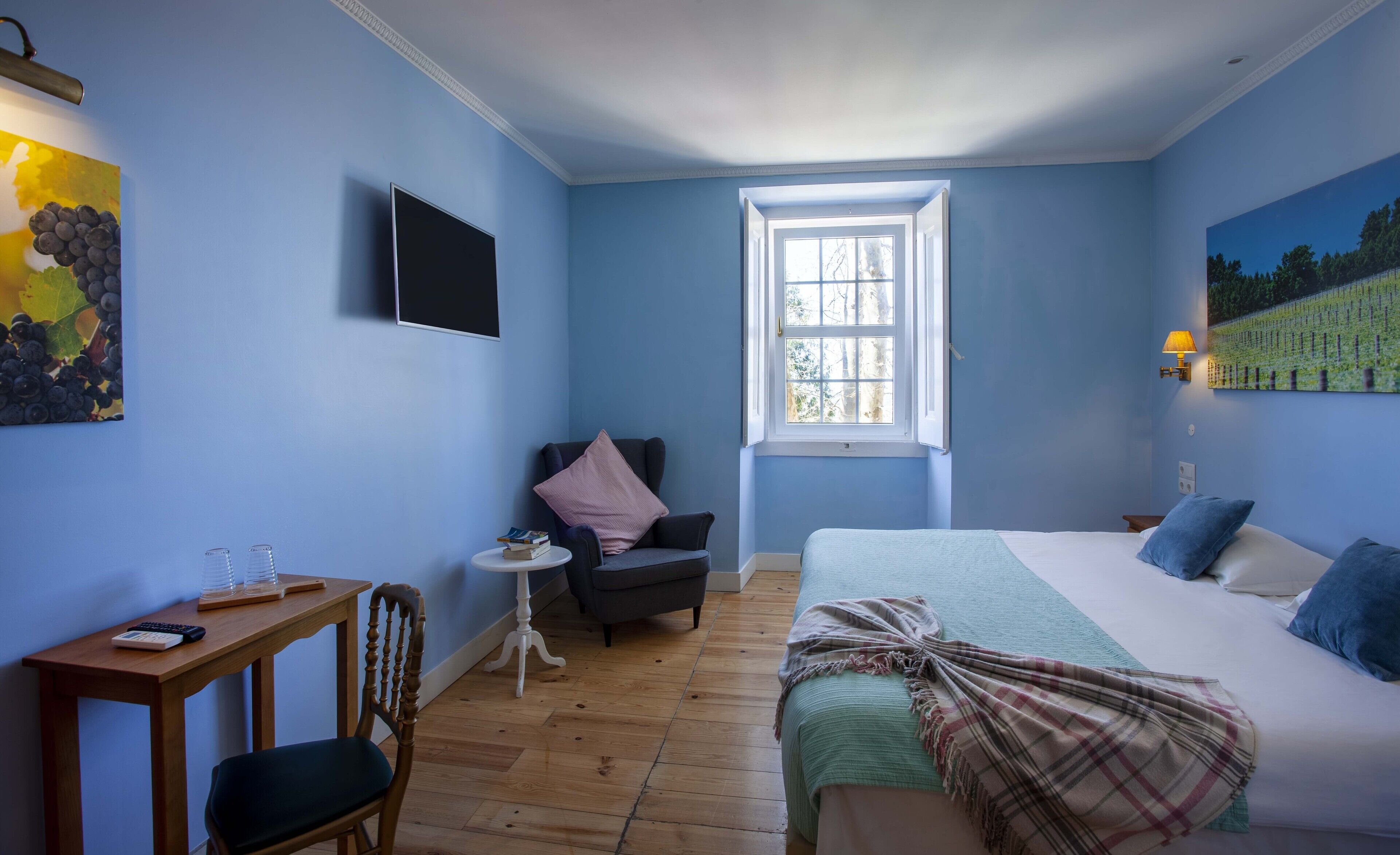 A room with a bed, a desk, a chair, a window, and artwork on the walls.