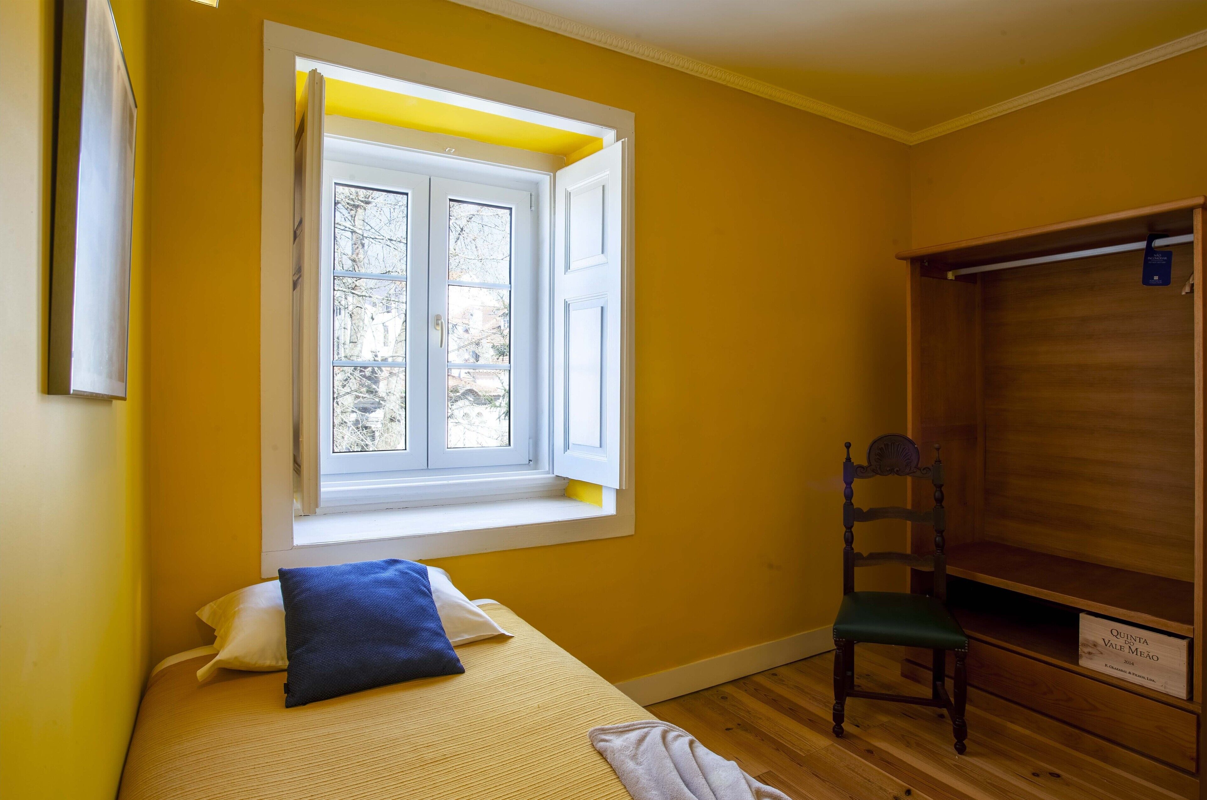 Une pièce avec un lit, une fenêtre dont les volets sont ouverts, une chaise en bois et une armoire.