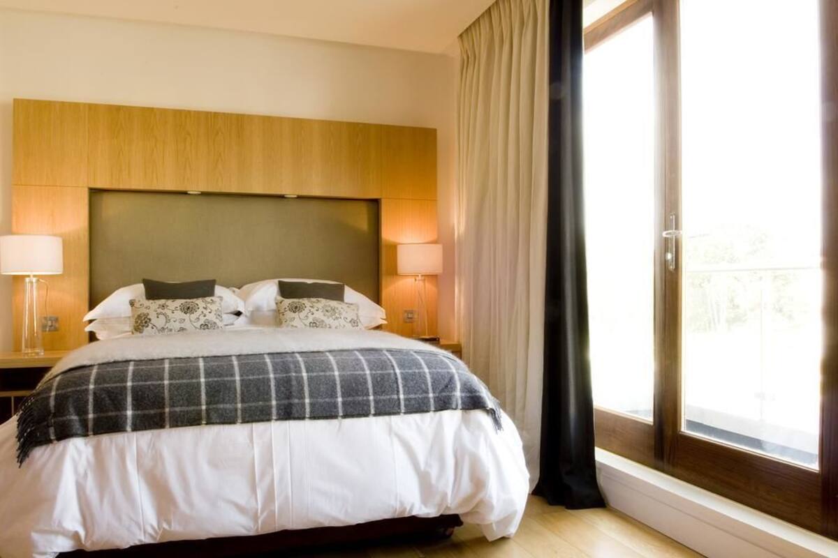 A hotel room with a large bed, two bedside lamps, a wooden headboard, and a view through a large window.