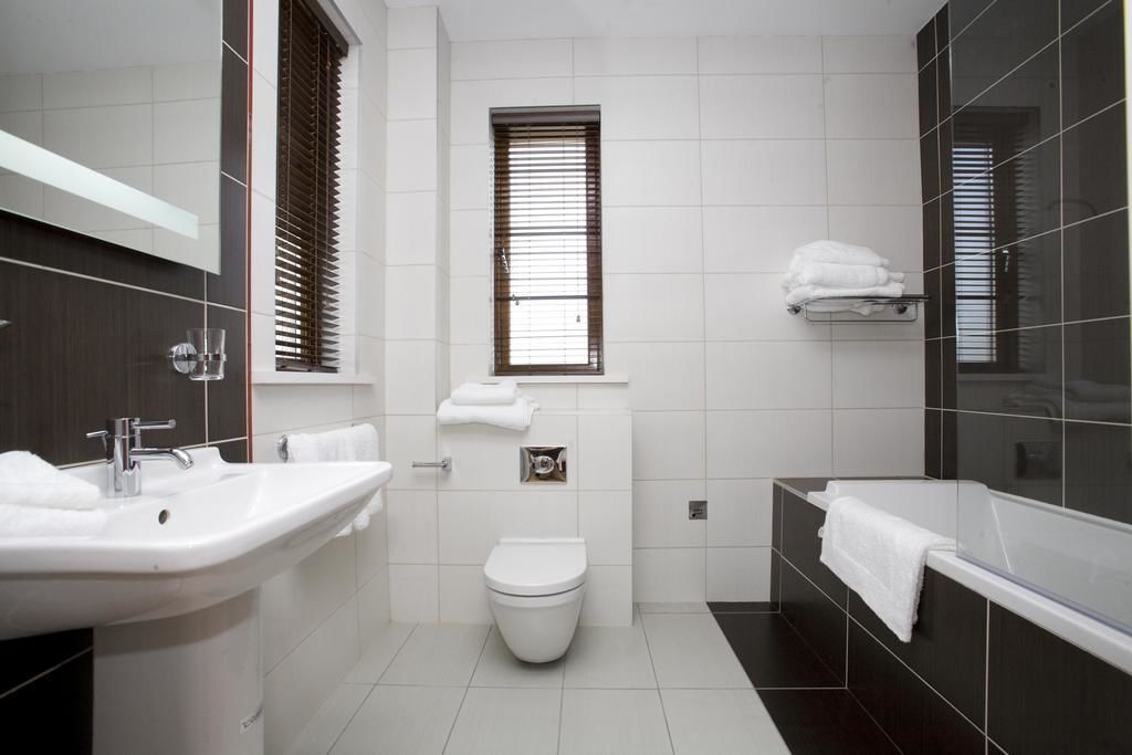 Un baño moderno con lavamanos, inodoro y bañera blancos, con baldosas oscuras y una ventana con persianas.