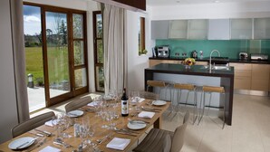 A modern kitchen with a central island, a dining area with a large table set for six, and a view of a green landscape through the windows. - Waterford Castle Lodges (Waterford)
