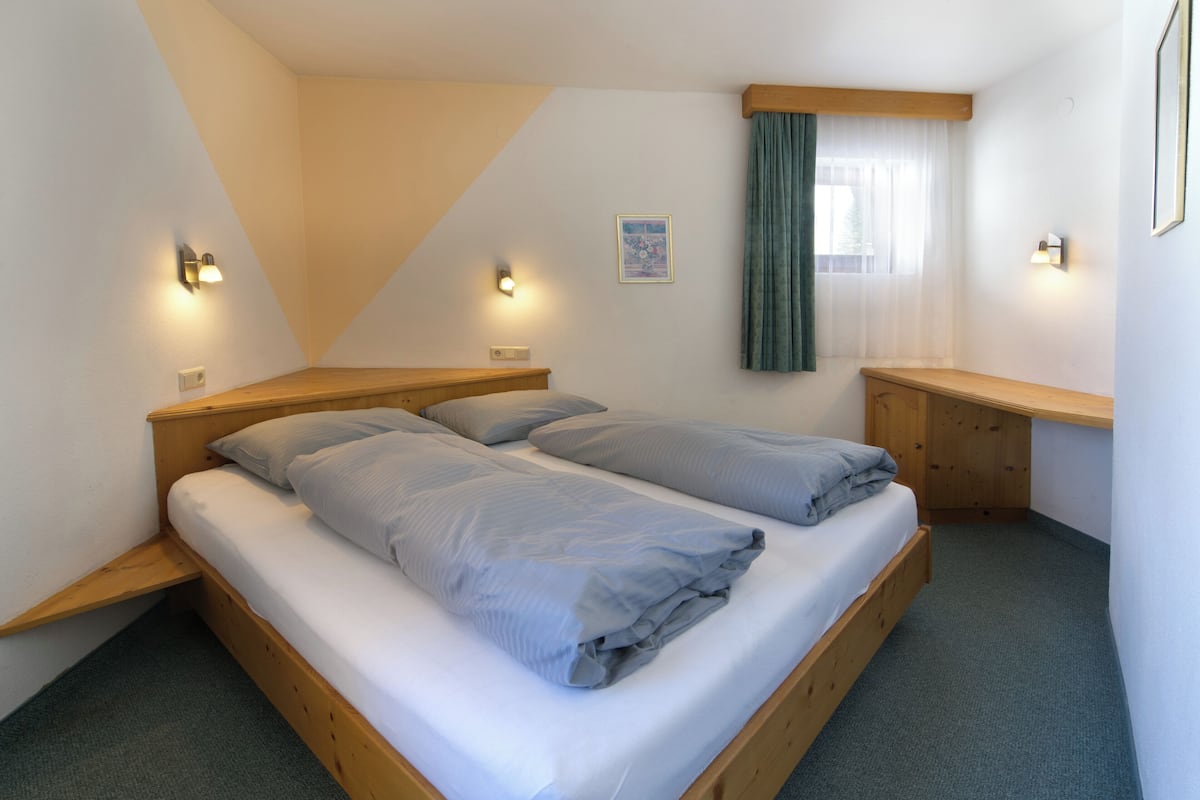 A small, well-lit bedroom with two single beds, a wooden bench, and a window with curtains.