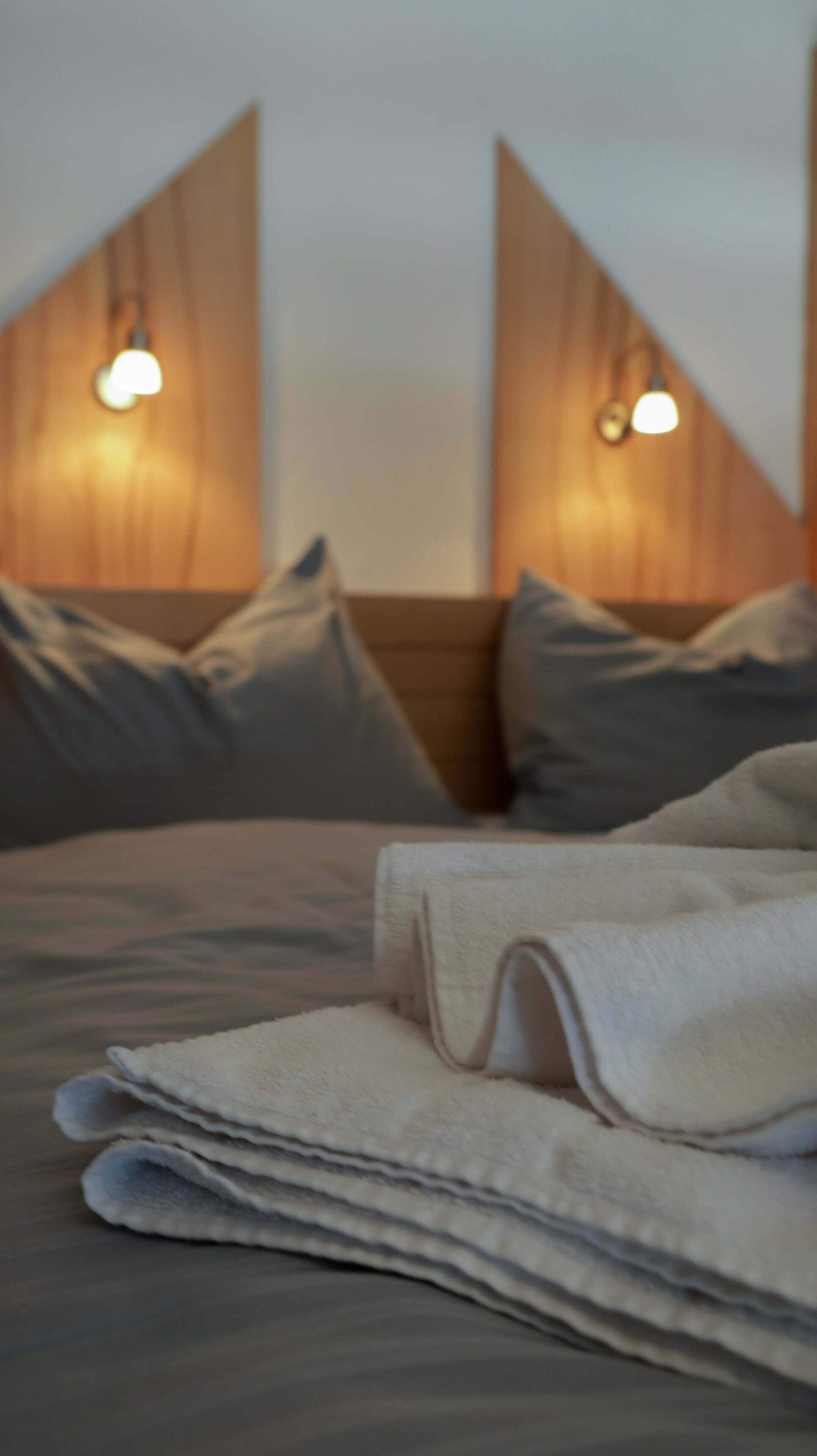 A neatly made bed with a folded towel, two pillows, and a headboard with wall-mounted lights.