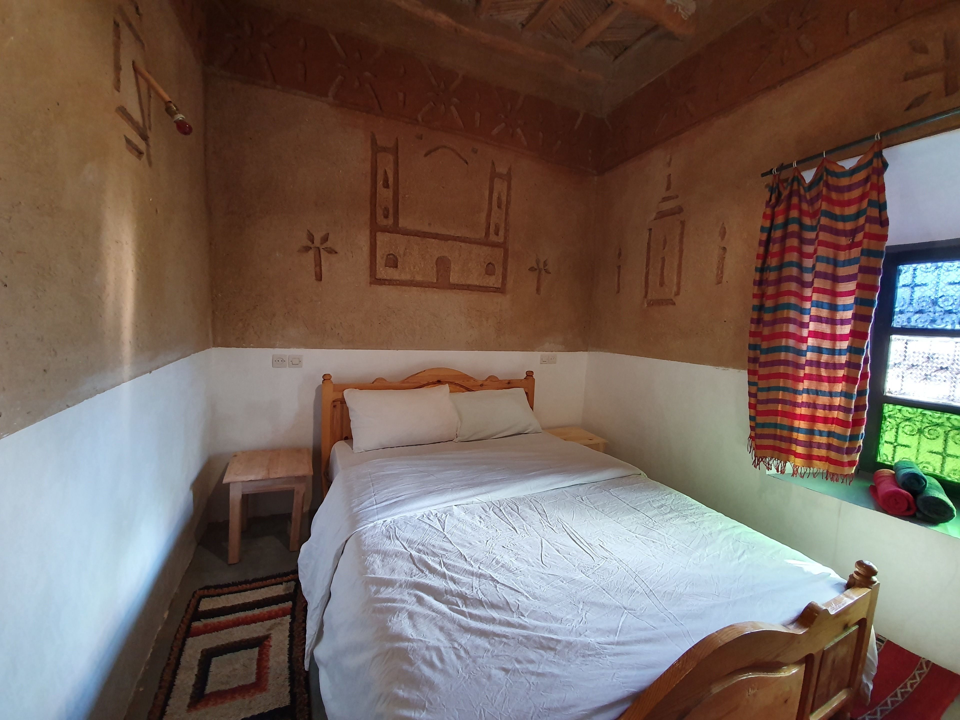 A bedroom with a wooden bed, a small bedside table, a window with a striped curtain, and a wall with decorative carvings.