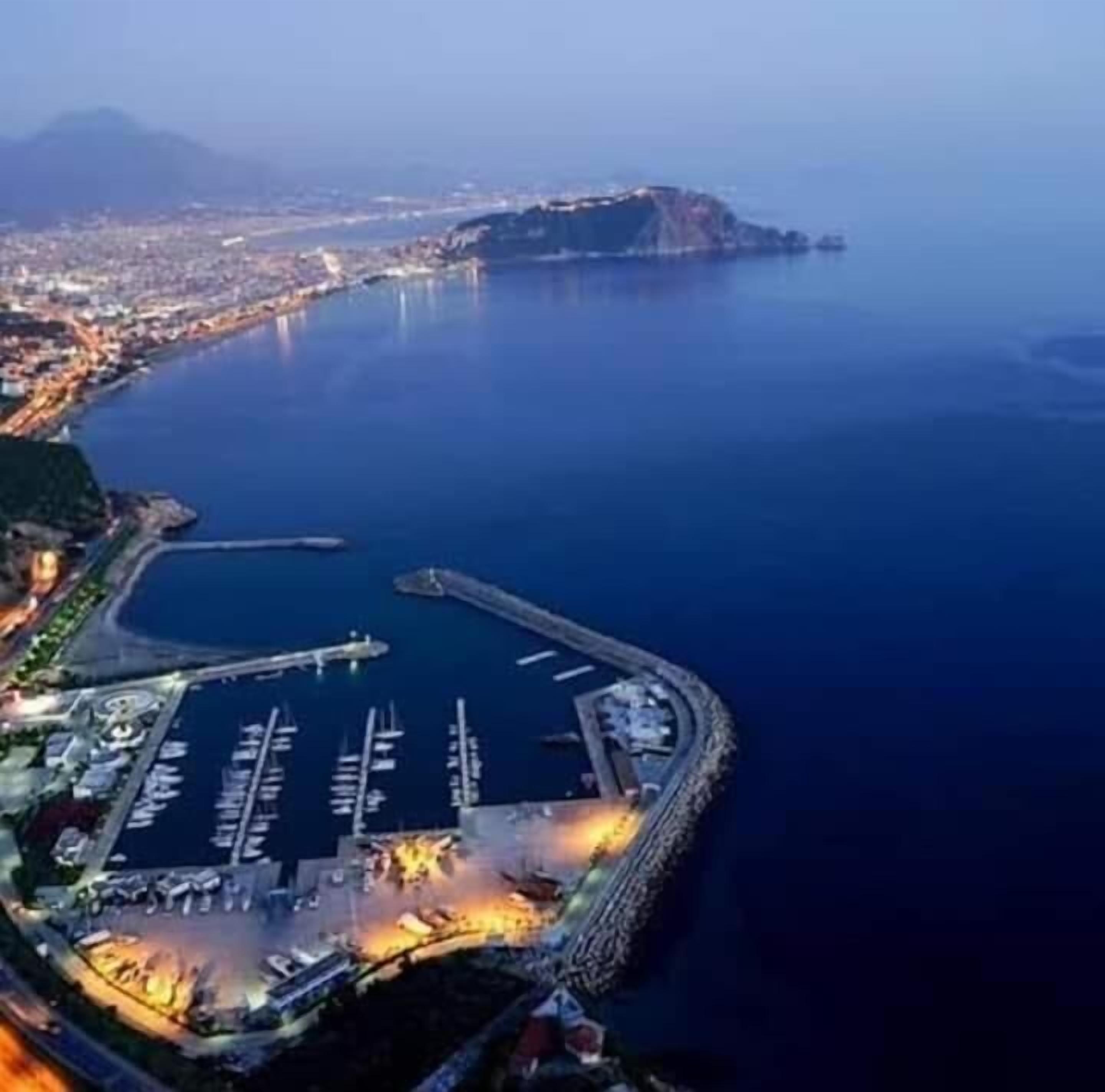 An aerial view of a coastal marina with boats docked, a large open water area, and a distant mountain range.