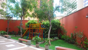 A courtyard with a playground, benches, potted plants, and a red wall. - Altavista Apartments (Fortaleza)