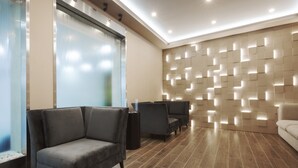 Un área de espera moderna con sofás grises, un panel de vidrio y una pared con paneles iluminados.
