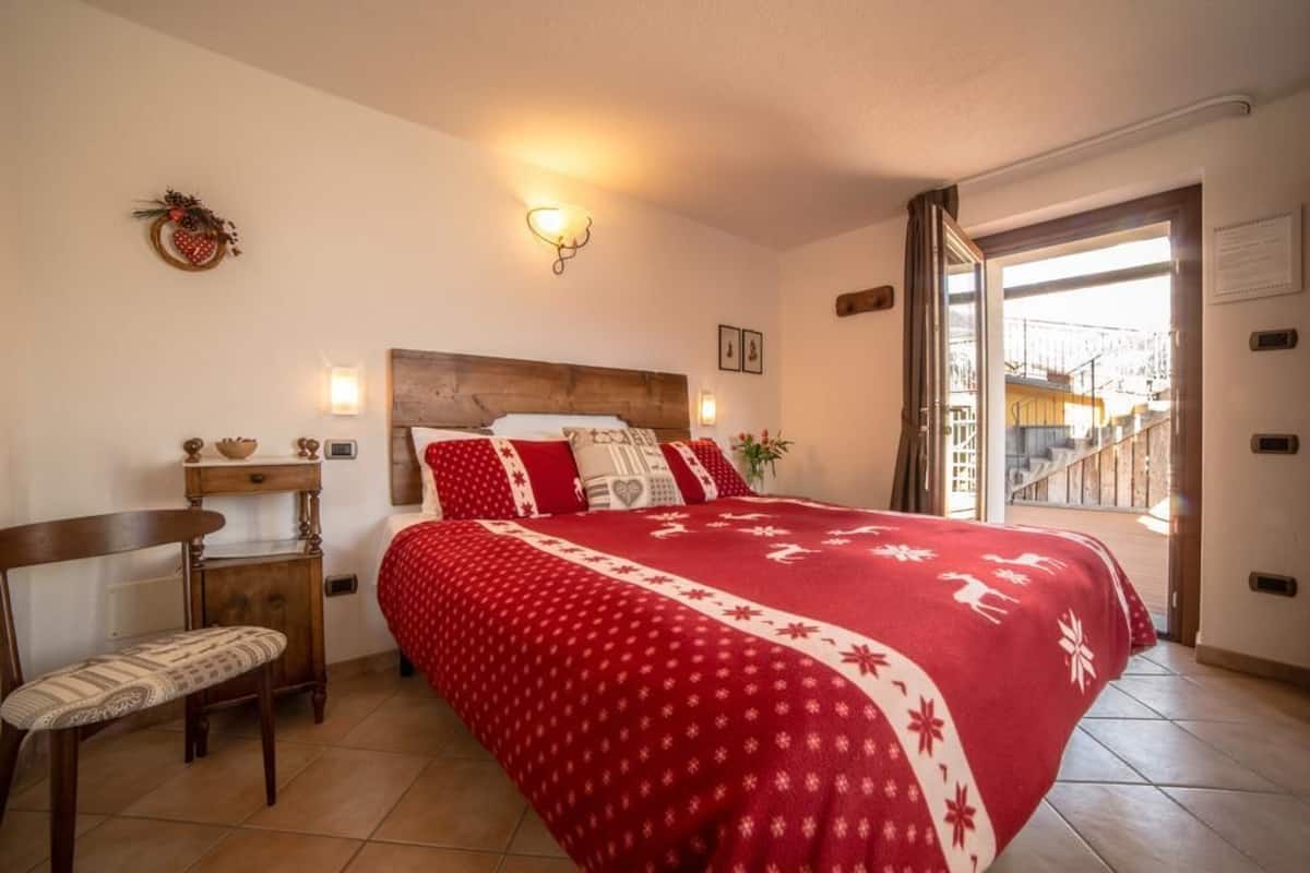 A bedroom with a wooden headboard, a red patterned bedspread, a wooden nightstand, a chair, and a view of a staircase through an open door.