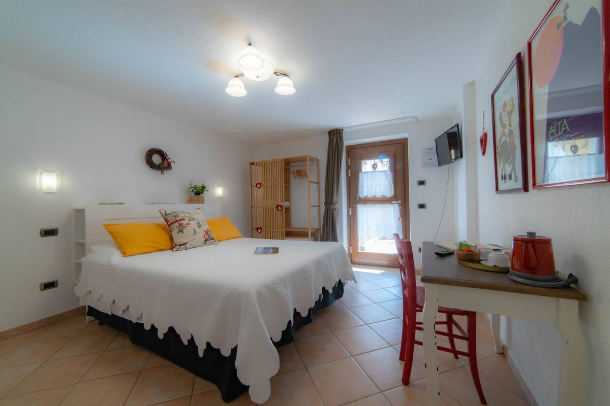 A bedroom with a bed, a desk, and a chair. There is a window with curtains, a door, and a wall-mounted TV.