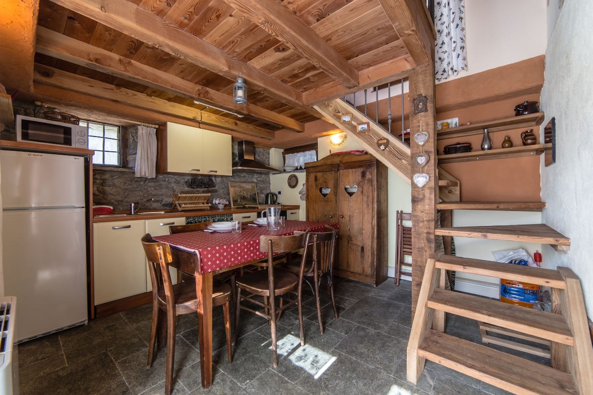 A rustic kitchen with wooden beams, a stone wall, a wooden dining table with chairs, and a wooden staircase with heart-shaped decorations.