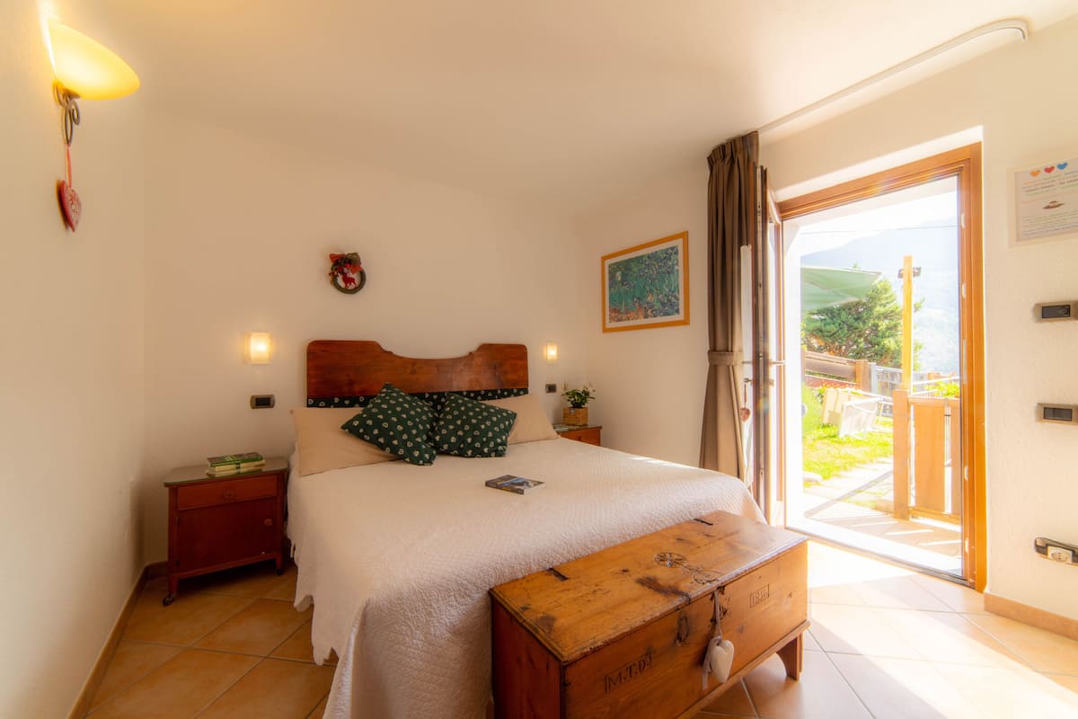 A bedroom with a bed, bedside tables, a wooden chest, a window with curtains, and a view of the outdoors.