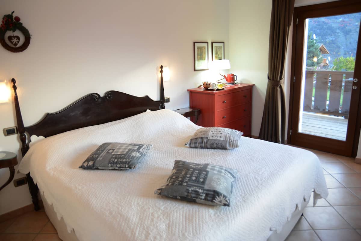 A neatly made bed with a white duvet and patterned pillows, a wooden headboard, a red nightstand with a lamp, and a view of a balcony with a wooden railing.