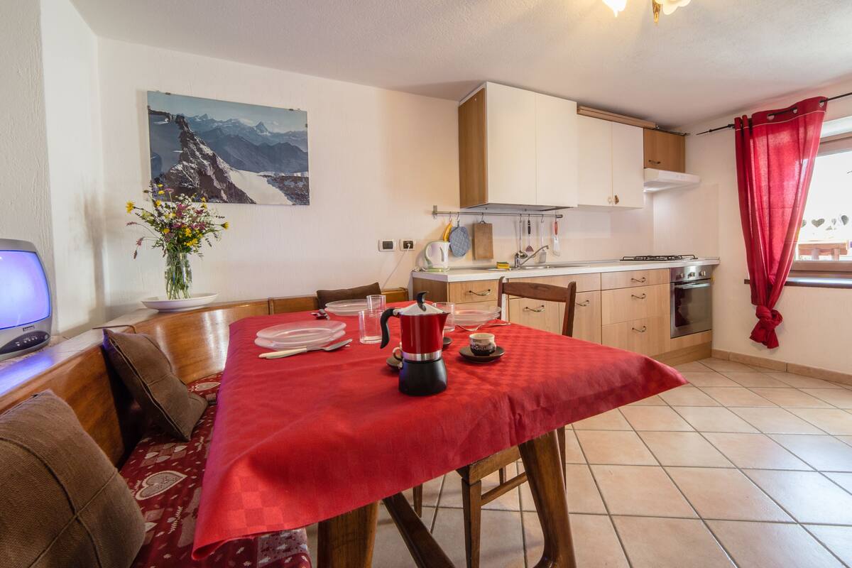A dining area with a red tablecloth, wooden chairs, a coffee pot, and a painting of mountains on the wall.