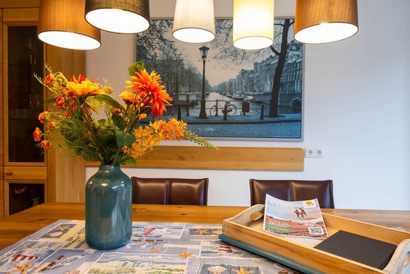 Un coin repas avec une table dressée pour deux personnes, un vase rempli de fleurs oranges, un menu imprimé et, au-dessus, un tableau représentant un paysage urbain.