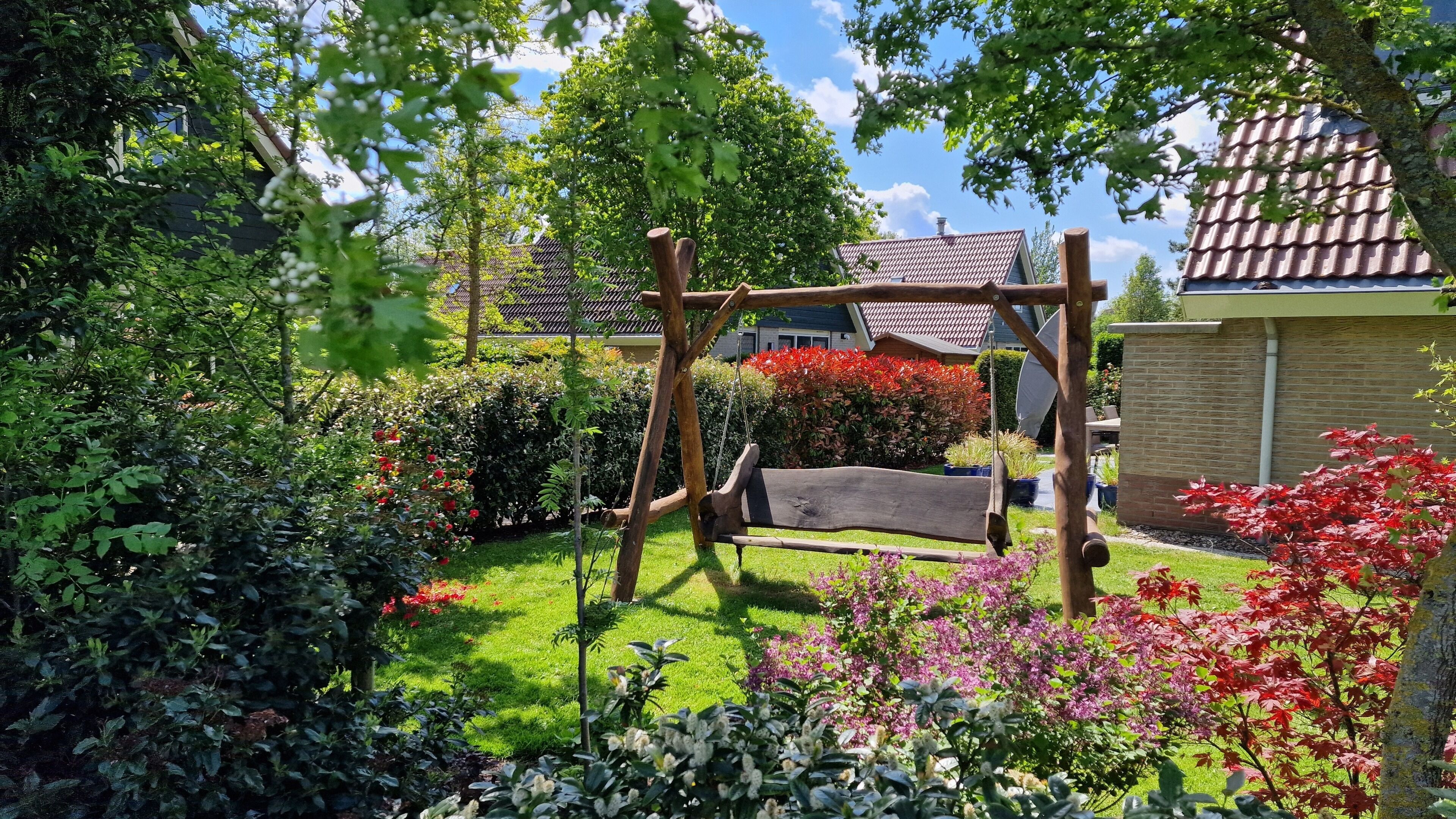 A garden with a wooden swing, lush greenery, and a house with a tiled roof.