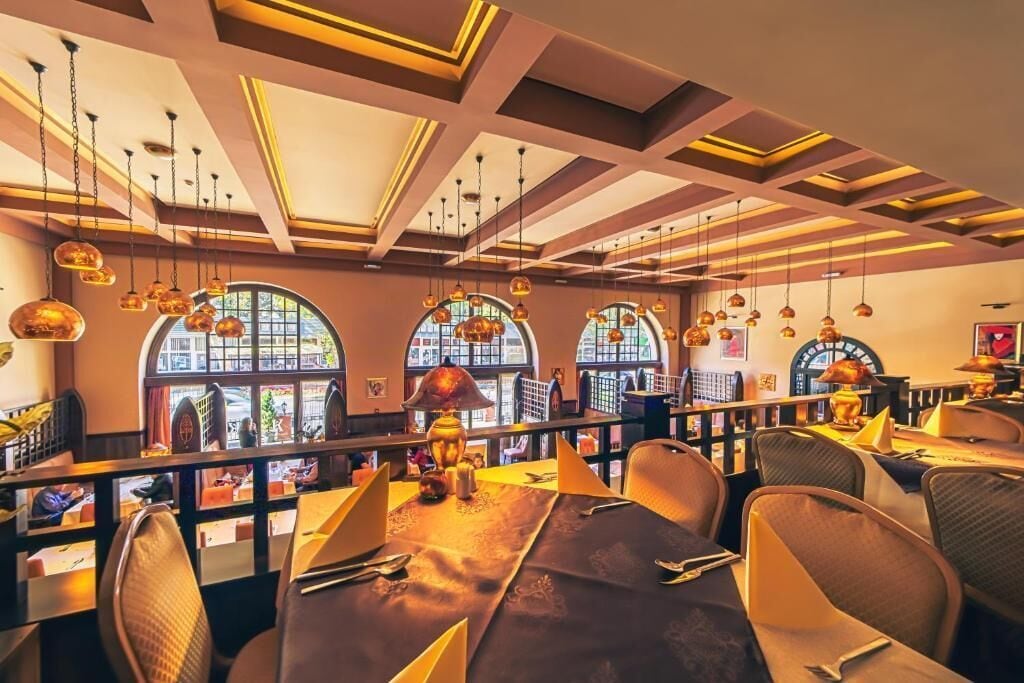 L’intérieur d’un restaurant se caractérise par de hauts plafonds, des fenêtres en arc de cercle, des luminaires suspendus et une longue table à manger.