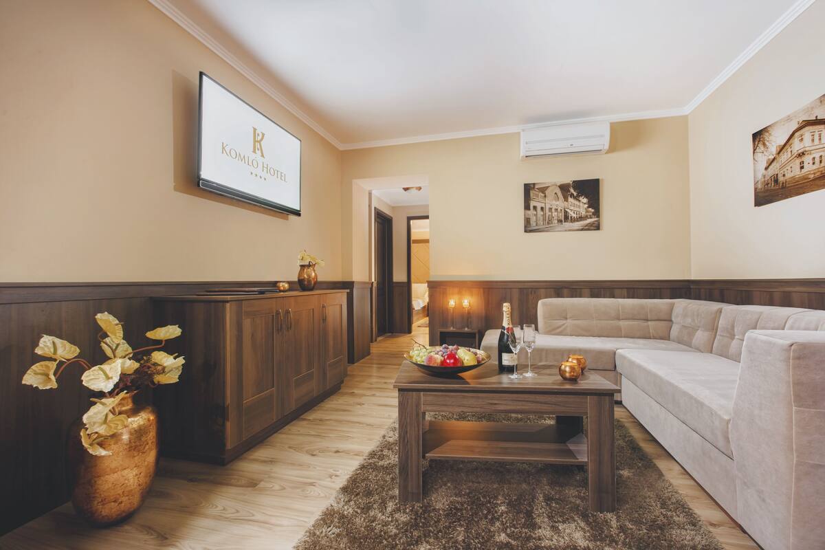 Un moderno vestíbulo de hotel con un mostrador de recepción de madera, un sofá, una mesa baja con frutero y copas de champán, y un cartel enmarcado sobre el mostrador.