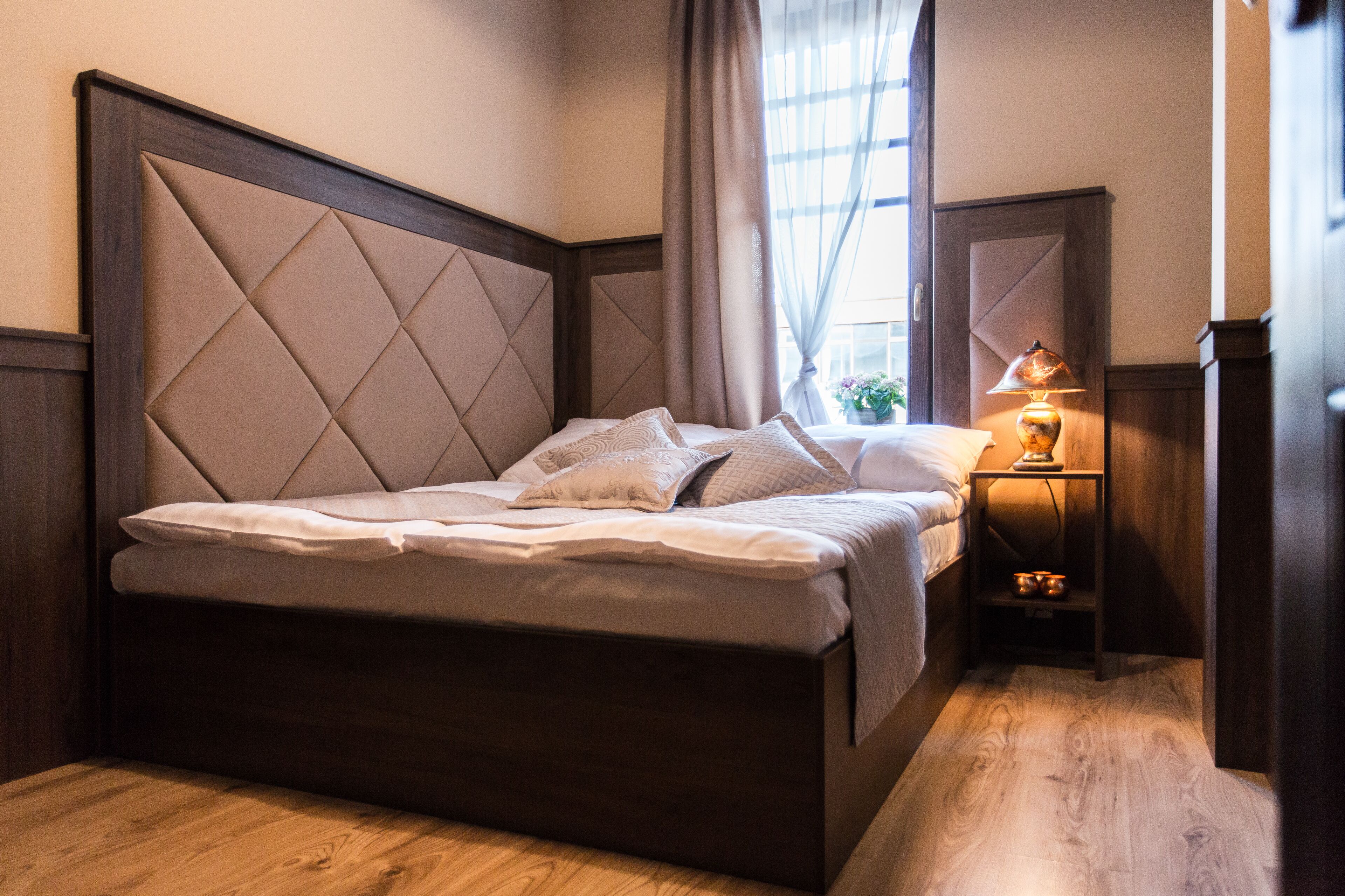 Una cama bien hecha con cabecera de madera, una mesita de noche con lámpara y una ventana con cortinas.