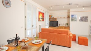 A modern living room with an orange sofa, a dining area with a table set for two, and a TV in the background. - Il Cedro (Salò)
