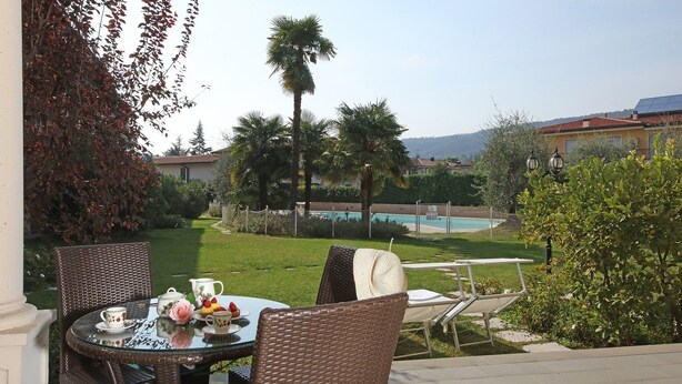 An outdoor seating area with wicker chairs and a glass table, overlooking a swimming pool and lush greenery. - Il Cedro (Salò)