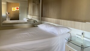 Ein Hotelzimmer mit einem Bett, einem Nachttisch aus Glas, einem Spiegel und einem an der Wand befestigten Telefon.