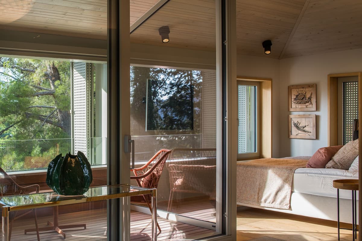 Un interior moderno con un vidrio que da a un balcón, una silla de mimbre, una mesa de vidrio y una cama con vista a los árboles.