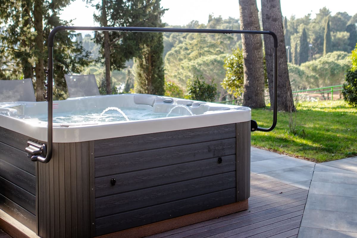 Un jacuzzi en una terraza de madera con barandilla metálica, rodeado de árboles altos y un espacio cubierto de césped.