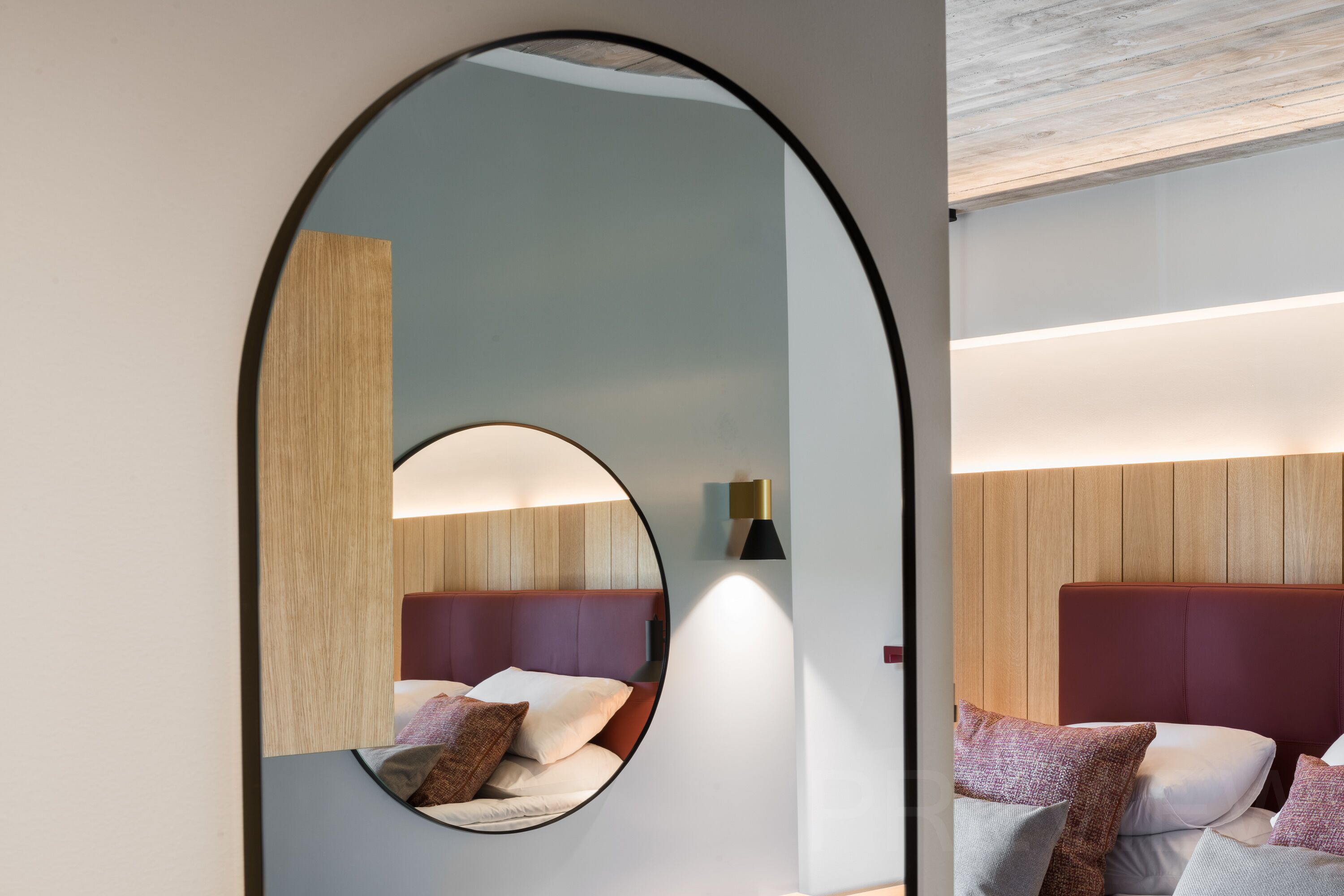 A modern hotel room with a circular mirror, a bed with a wooden headboard, and a wall-mounted light fixture.