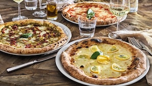 Una mesa de restaurante con tres pizzas, copas de vino y una botella de agua.