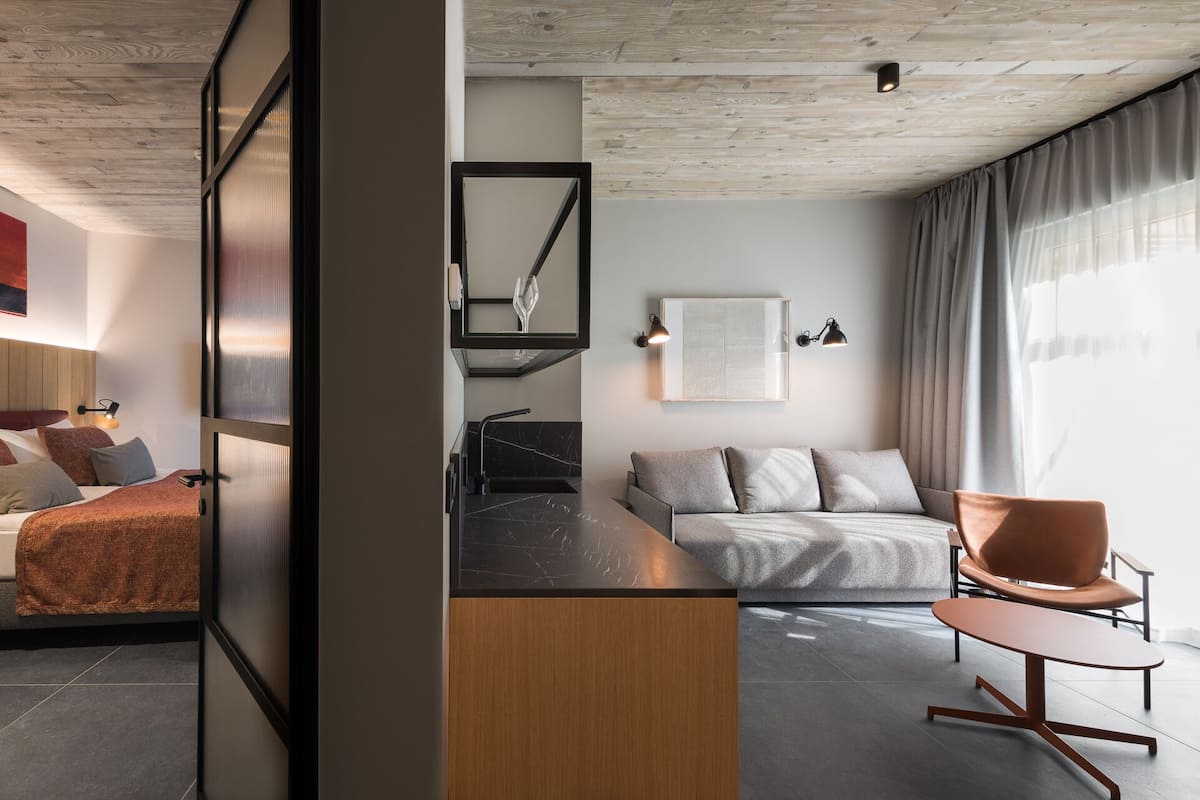 Una habitación de hotel moderna con cama, sofá, sillón, escritorio y una ventana grande con cortinas.