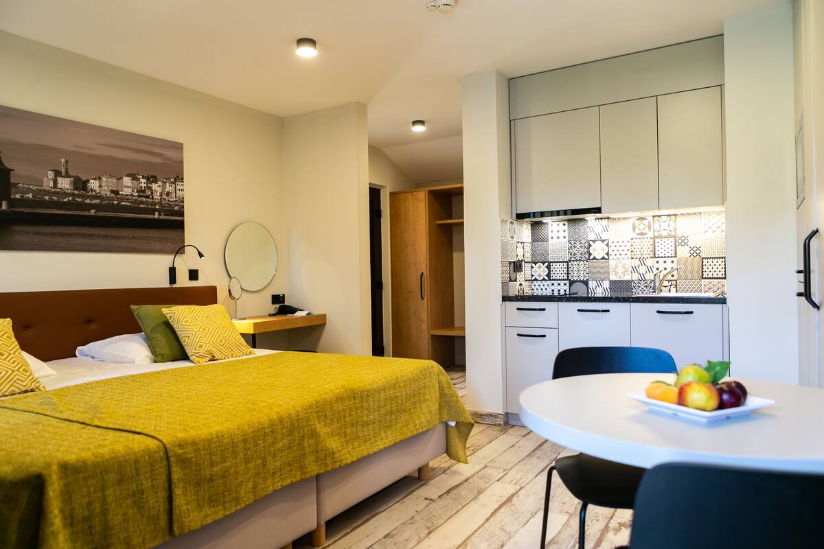 Una habitación de hotel con una cama, una mesita de noche con lámpara, un espejo, una mesa de comedor con fruta y una cocina con armarios y salpicadero de azulejos.