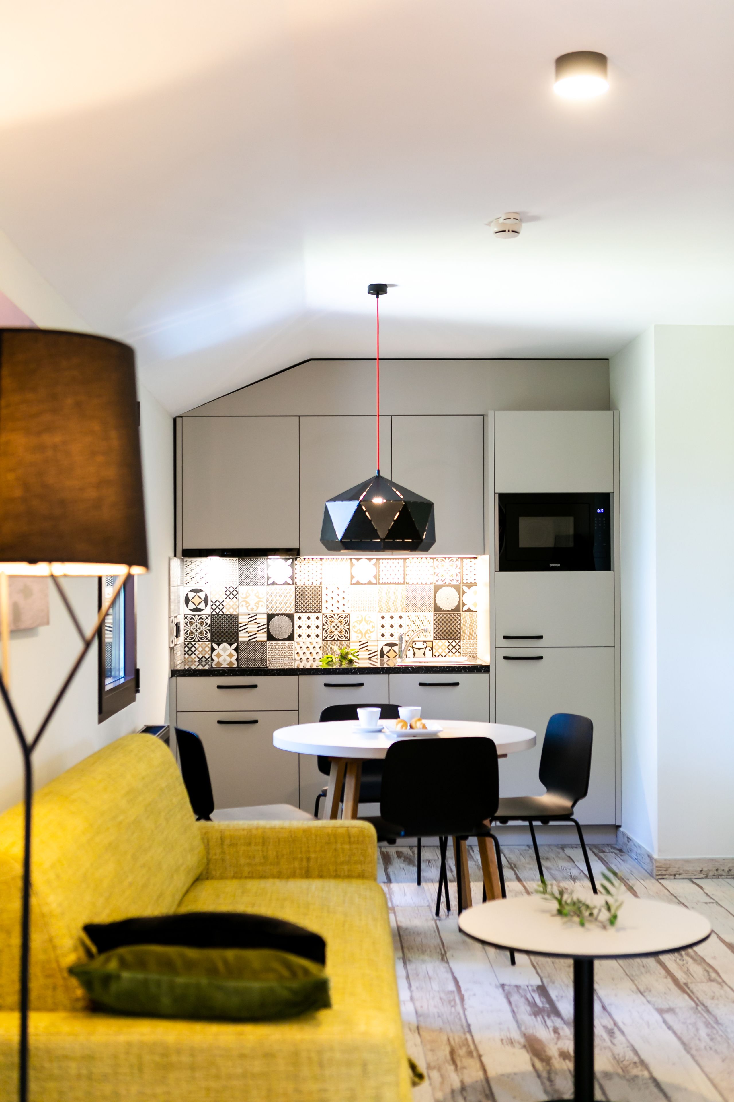 Et moderne køkken med et geometrisk bagpanel, et spiseområde med et rundt bord og stole, samt en gul sofa med pude.