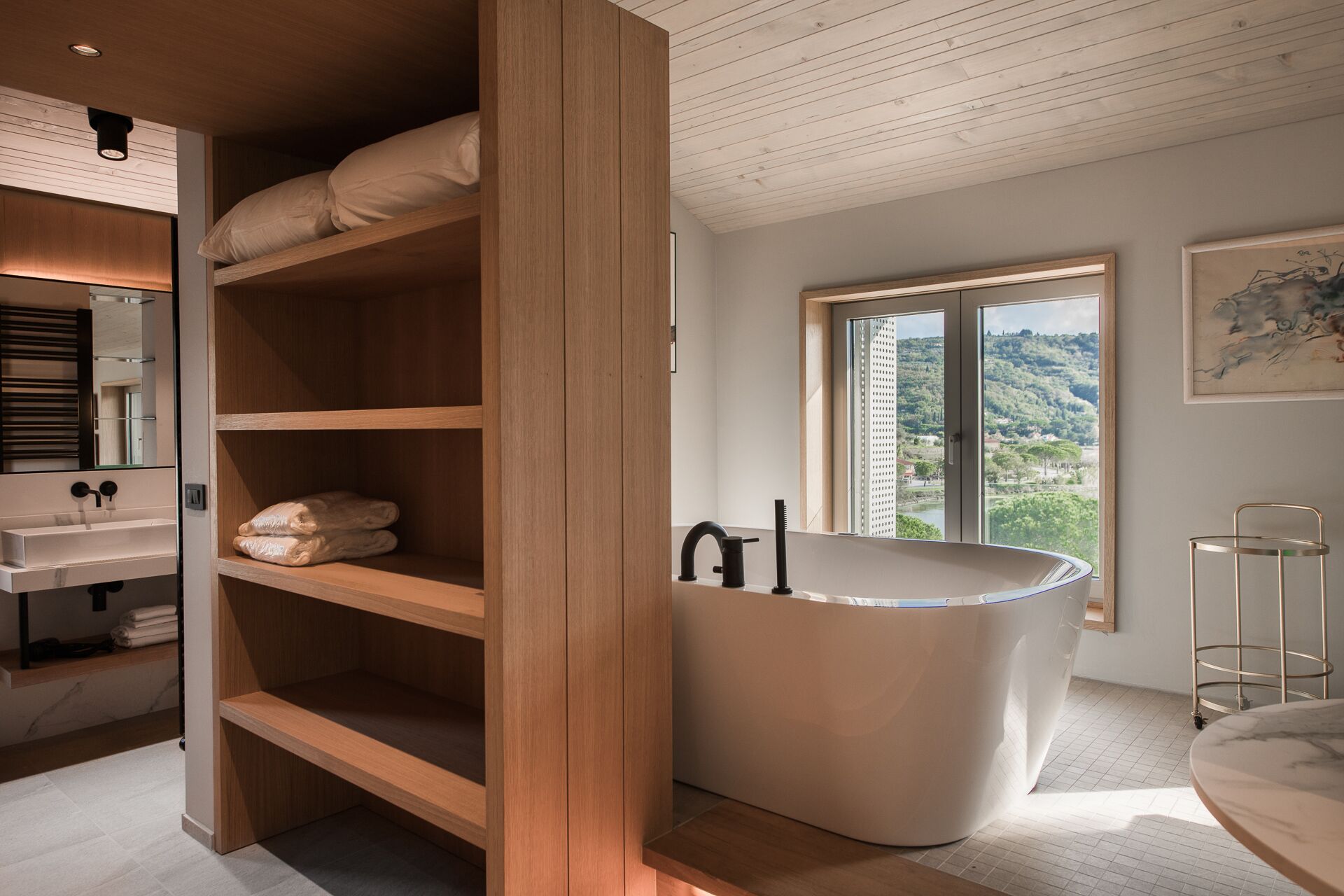 A modern bathroom with a freestanding bathtub, wooden shelving, and a large window offering a scenic view.