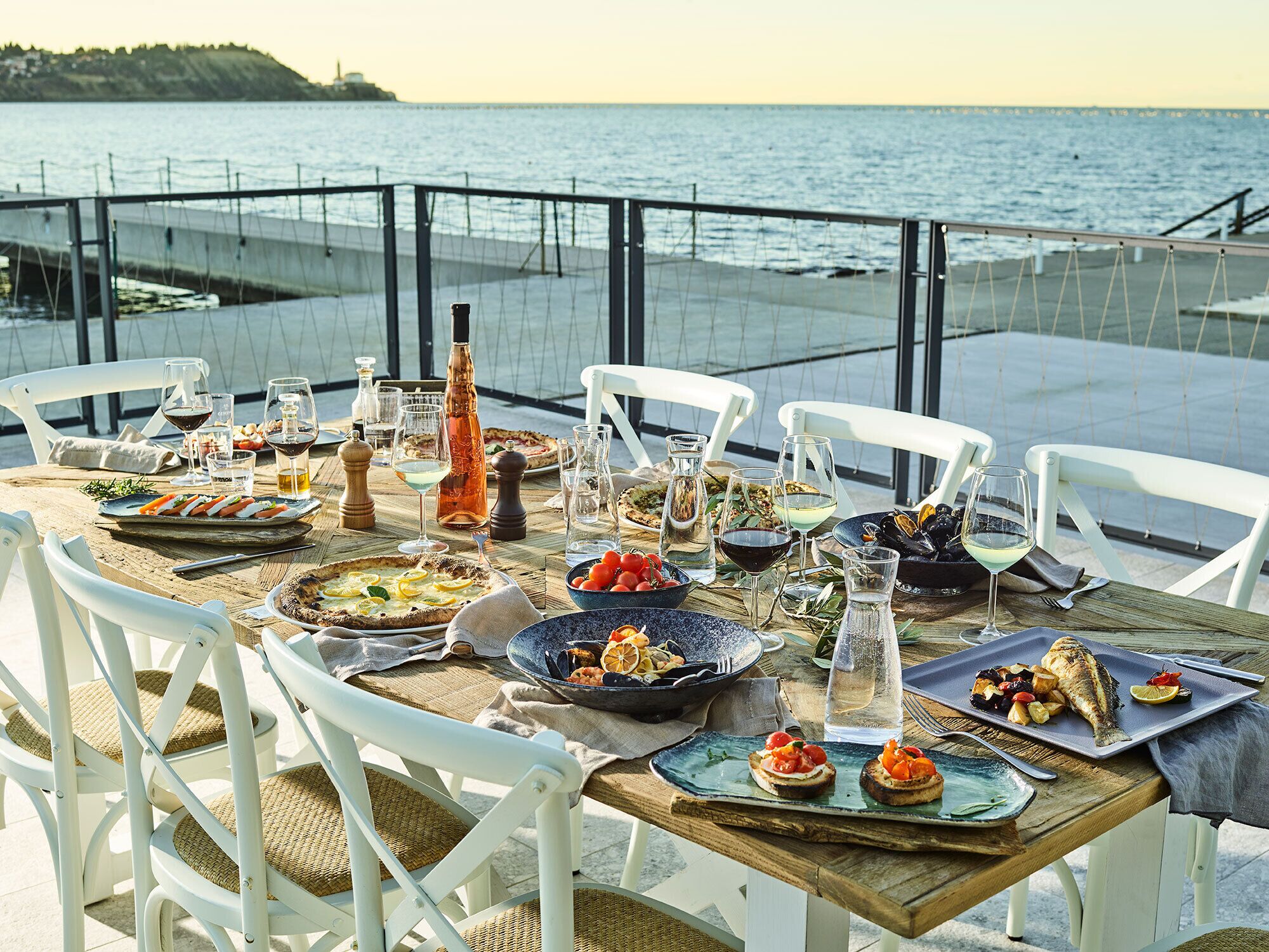 A table set for a meal with various dishes and drinks, overlooking a body of water.