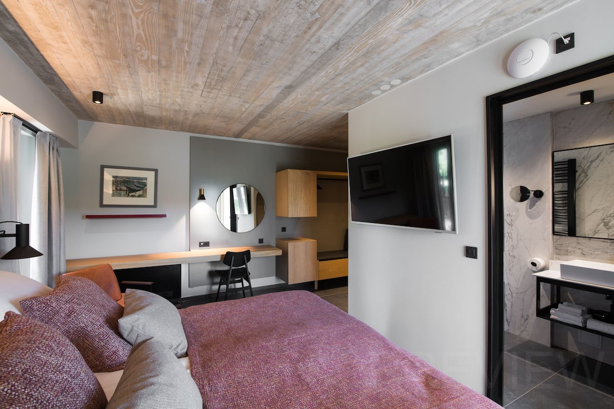 Una habitación de hotel moderna con techo de madera, cama con ropa de cama estampada, escritorio con silla, televisor montado en la pared y baño con lavamanos de mármol.