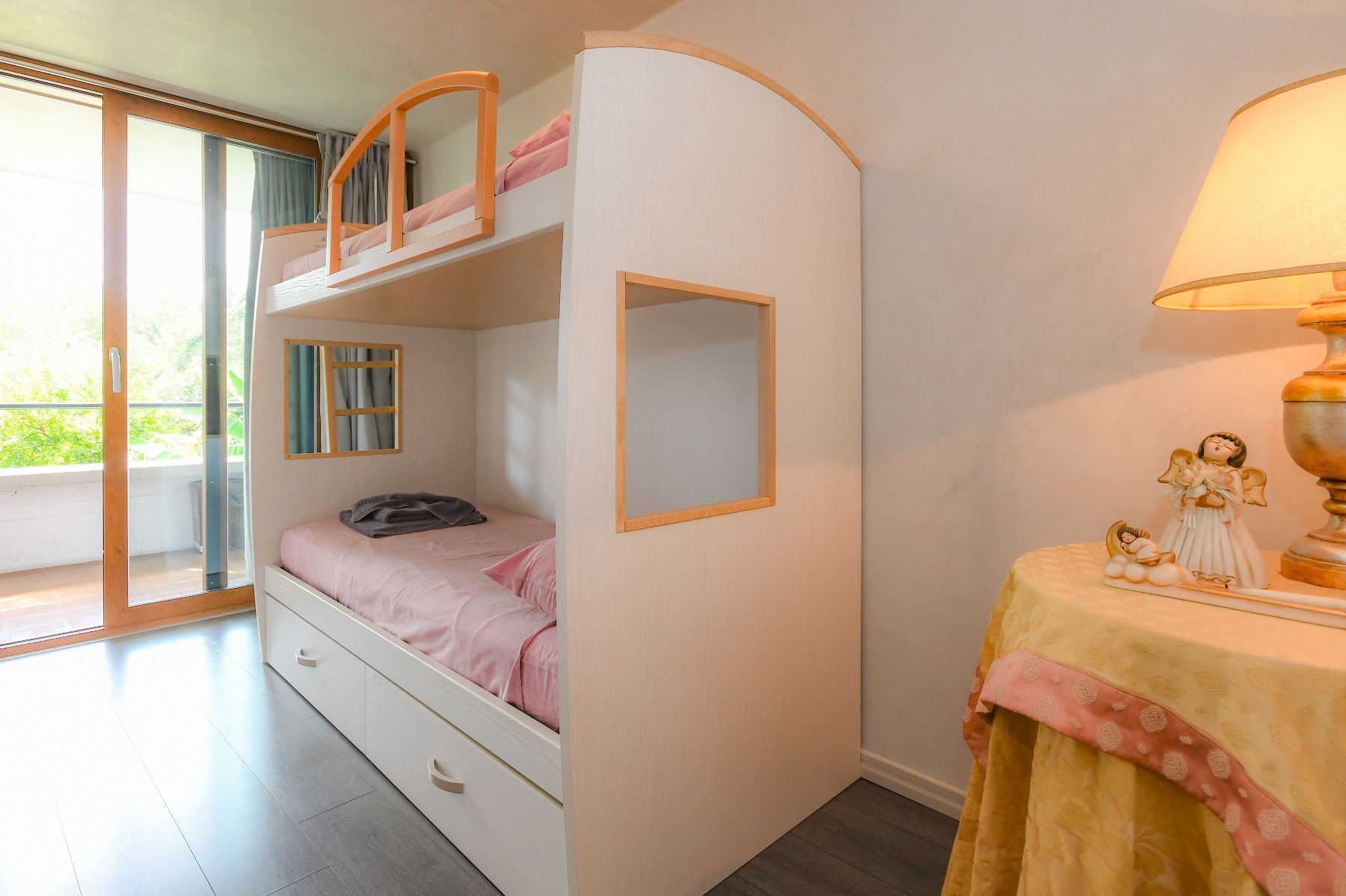 A bedroom with a bunk bed, a single bed, a small table with a lamp, and a window with curtains.