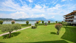 A well-maintained garden with a pathway, a building with balconies overlooking a lake, and mountains in the background. - Il Sogno del Lago (Manerba del Garda)