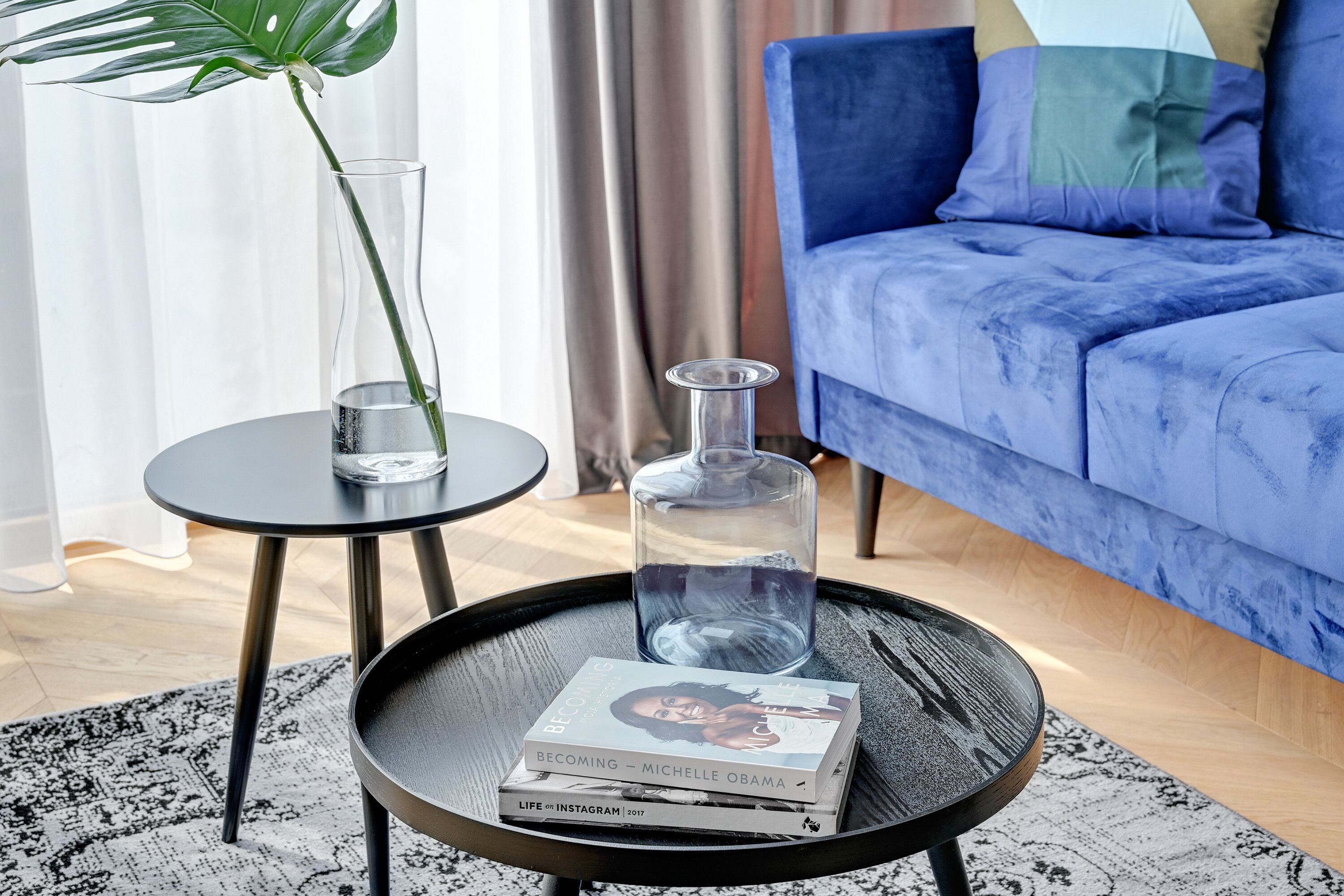 A modern living room with a blue sofa, a glass coffee table, a book titled 'Becoming Michelle Obama', and a potted plant.