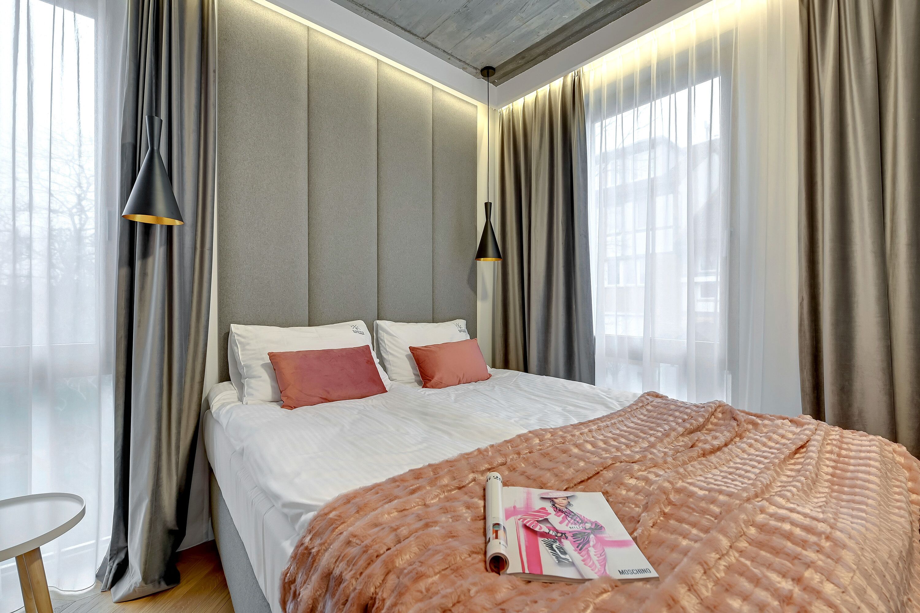 A modern hotel room with a large bed, grey padded headboard, and a magazine on the bed.