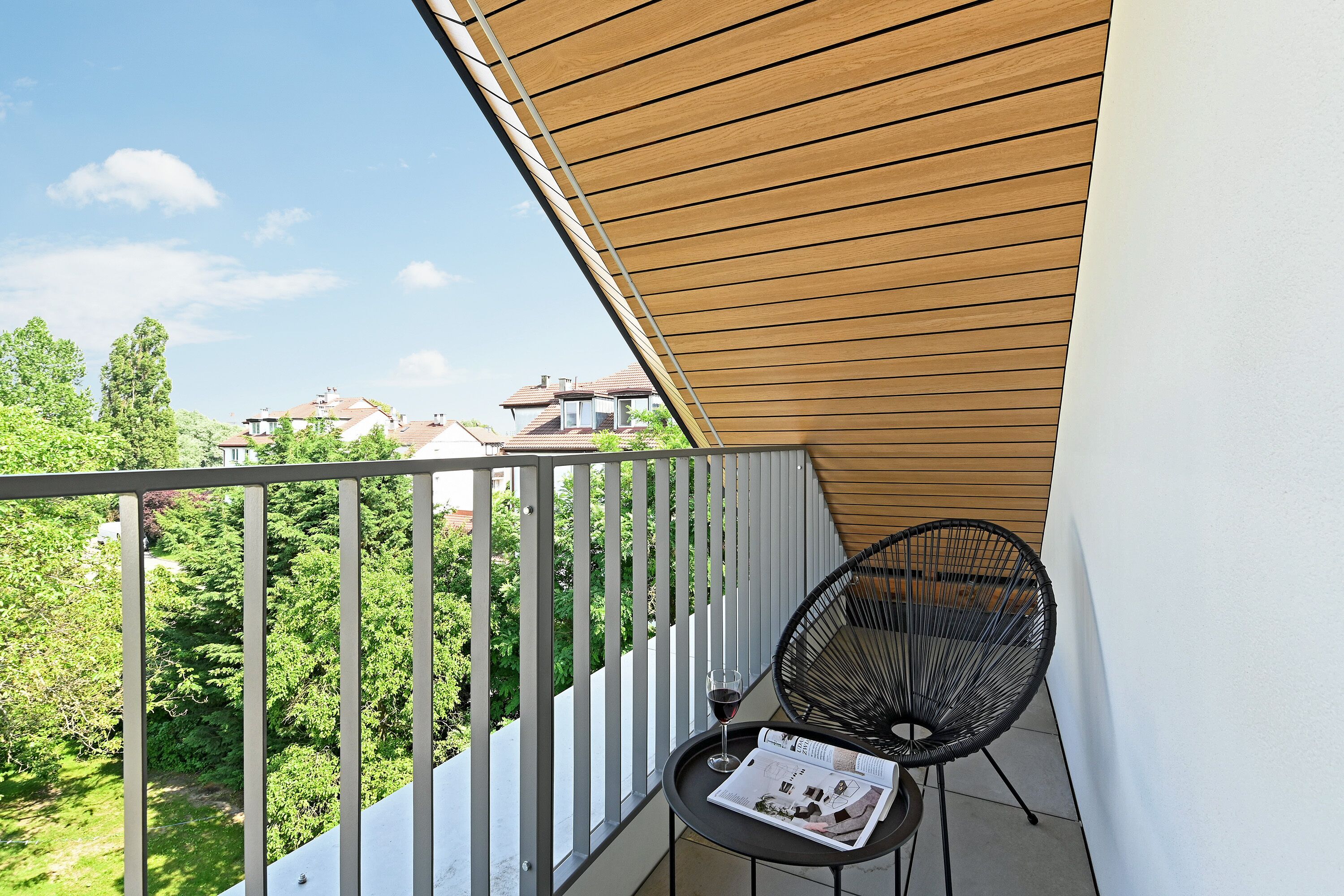 A balcony with a wooden ceiling, a metal railing, a black chair, and a glass of wine on a table with a magazine.