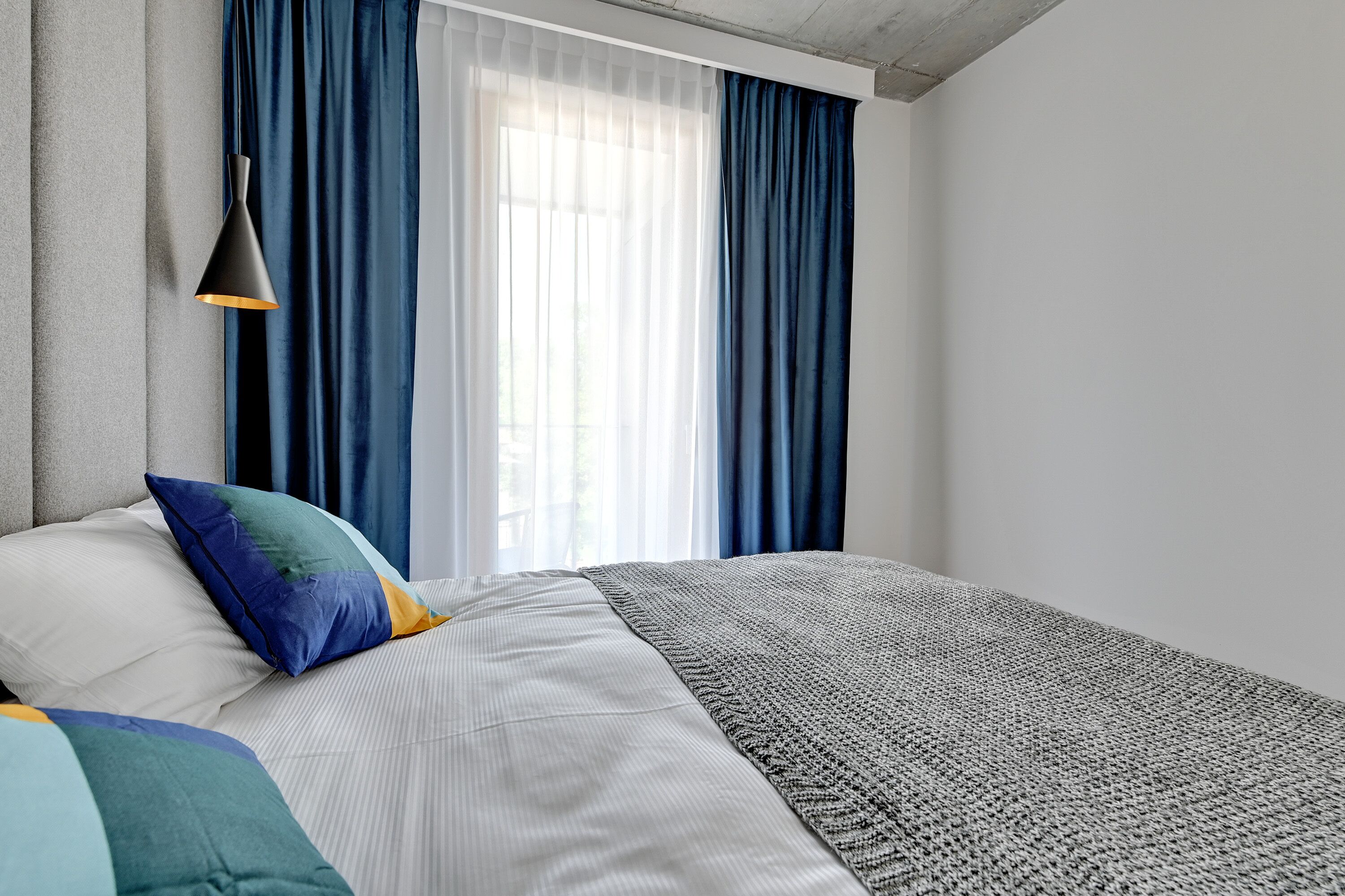 A bedroom with a bed, pillows, a blanket, a hanging lamp, and a window with curtains.